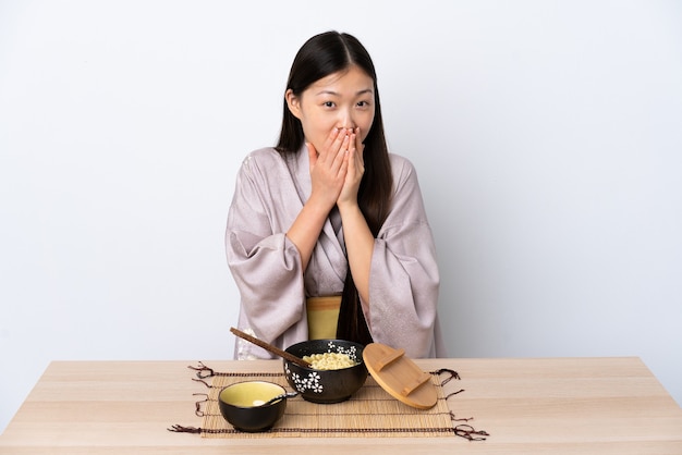 Jovem chinesa vestindo quimono e comendo macarrão feliz e sorridente, cobrindo a boca com as mãos