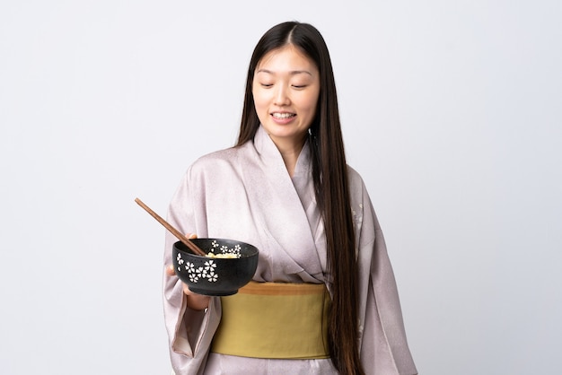 Jovem chinesa vestindo quimono sobre fundo branco isolado com uma expressão feliz enquanto segura uma tigela de macarrão com pauzinhos