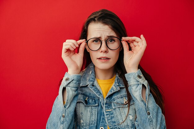 Jovem chocada mantém as mãos na borda dos óculos parece com suspiros de expressão omg de choque, ouve notícias incríveis, usa jaqueta jeans, isolada em um fundo vermelho com espaço de cópia