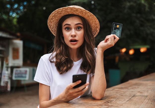 Jovem chocada segurando um cartão de crédito de plástico enquanto usa o telefone celular em um café ao ar livre