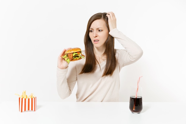 Jovem chocada sentada à mesa com hambúrguer, batata frita, coca-cola em frasco de vidro, isolado no fundo branco. Nutrição adequada ou fast food clássico americano. Área de publicidade com espaço de cópia.