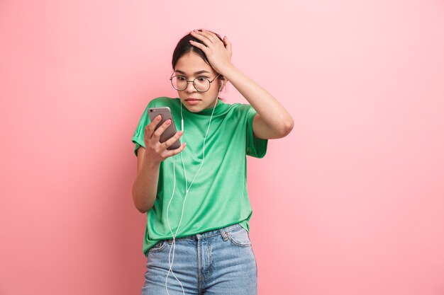 jovem chocada usando óculos redondos usando fones de ouvido enquanto segura o smartphone isolado sobre a parede rosa