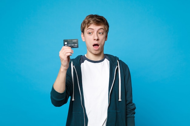 Jovem chocado em roupas casuais segurando, olhando no cartão do banco de crédito, mantendo a boca aberta isolada na parede azul. emoções sinceras de pessoas, conceito de estilo de vida.