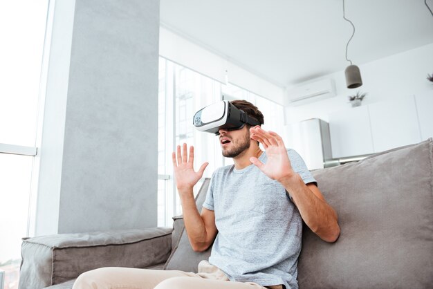 Jovem chocado usando um dispositivo de realidade virtual enquanto está sentado no sofá