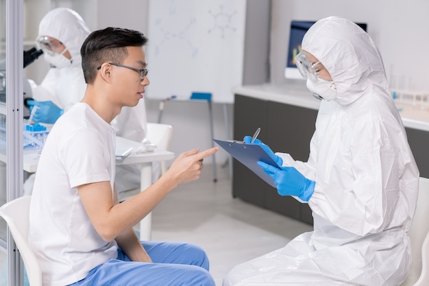 Jovem cientista com óculos de proteção, máscara e macacão fazendo anotações em um documento enquanto consulta um chinês no laboratório