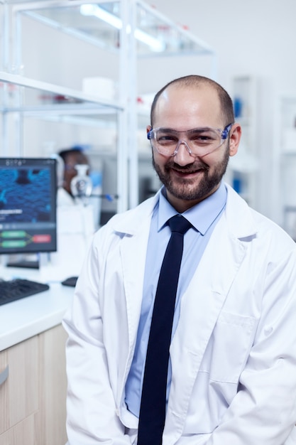 Jovem cientista da bioquímica usando óculos de proteção, sorrindo para a câmera. Grave especialista em genética em laboratório com tecnologia moderna para investigações médicas com assistente africano em segundo plano.