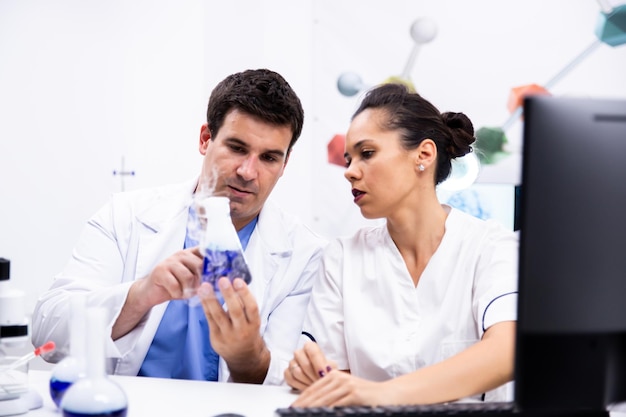 Jovem cientista de jaleco branco segurando uma garrafa com um líquido azul fumegante e explicando para seu assistente.
