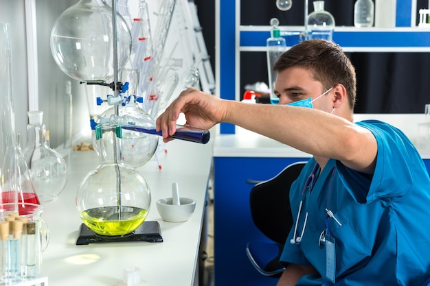 Jovem cientista masculino de uniforme usando uma máscara está segurando um grande tubo de ensaio com um líquido azul e fazendo algumas pesquisas em um laboratório. Conceito de saúde e biotecnologia