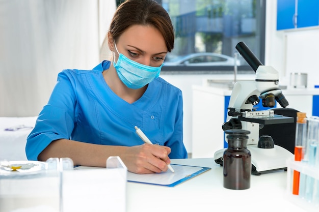 Jovem cientista mulher atraente em uniforme escrevendo notas de sua pesquisa em um laboratório. conceito de saúde e biotecnologia