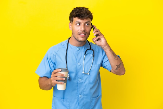 Jovem cirurgião médico homem isolado em um fundo amarelo segurando um café para levar e um celular