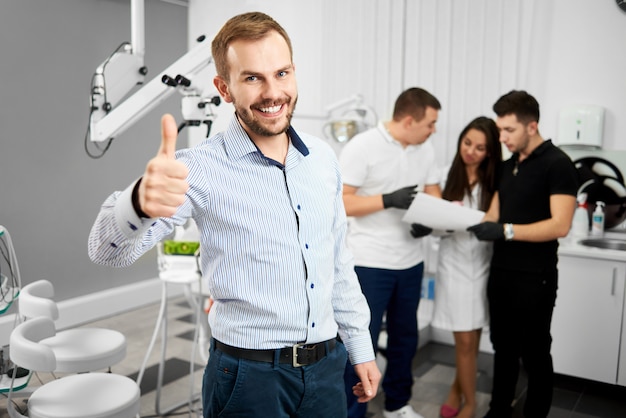 Jovem cliente masculino atraente de uma odontologia está sorrindo para a câmera e mostrando polegares para cima sendo feliz após o tratamento. Três especialistas em odontologia estão discutindo algo em segundo plano.