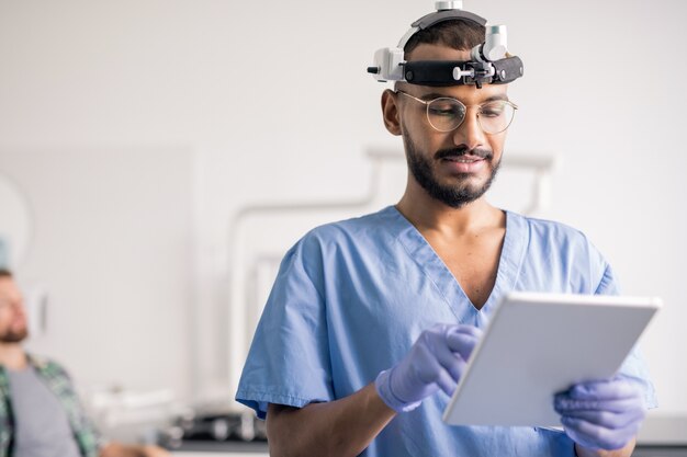 Jovem clínico de uniforme e equipamento médico na cabeça usando o touchpad enquanto rola os dados online no hospital