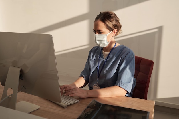 Jovem clínico sério em máscara e uniforme inserindo dados médicos no computador