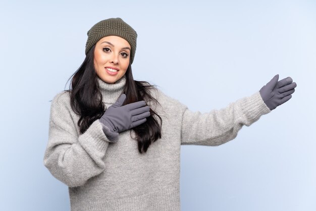 Foto jovem colombiana com chapéu de inverno sobre parede azul, estendendo as mãos para o lado para convidar para vir