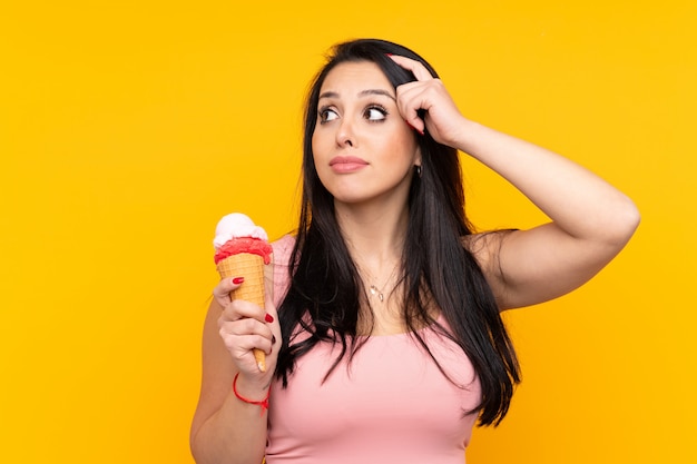 Jovem colombiana segurando um sorvete de corneta sobre parede amarela isolada, tendo dúvidas e com a expressão do rosto confuso