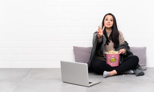 Jovem colombiana segurando uma tigela de pipocas e mostrando um filme em um laptop feliz e contando três com os dedos