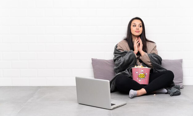 Jovem colombiana segurando uma tigela de pipocas e mostrando um filme em um laptop planejando algo