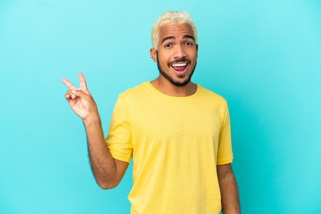 Jovem colombiano bonito isolado em um fundo azul sorrindo e mostrando sinal de vitória