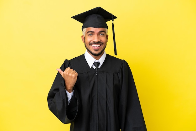 Jovem colombiano graduado pela universidade isolado em um fundo amarelo apontando para o lado para apresentar um produto