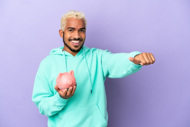 Jovem colombiano segurando um cofrinho isolado no fundo roxo e fazendo um gesto de polegar para cima