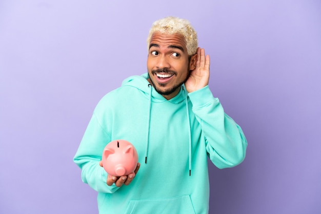 Jovem colombiano segurando um cofrinho isolado no fundo roxo, ouvindo algo colocando a mão na orelha