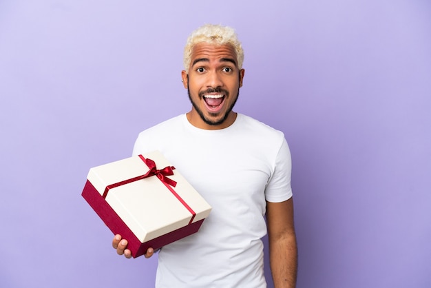 Jovem colombiano segurando um presente isolado em um fundo roxo com expressão facial surpresa