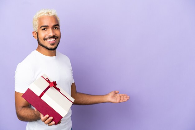 Jovem colombiano segurando um presente isolado em um fundo roxo, estendendo as mãos para o lado para um convite para vir