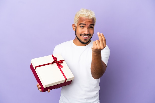 Jovem colombiano segurando um presente isolado no fundo roxo fazendo gesto de dinheiro