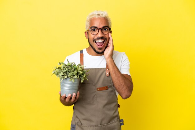 Jovem colombiano segurando uma planta isolada em um fundo amarelo com expressão facial de surpresa e choque