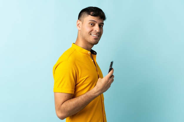 Jovem colombiano usando celular isolado em azul sorrindo