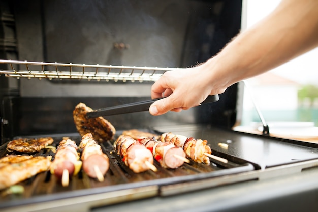 Jovem com as mãos virando a carne com um garfo na churrasqueira a gás