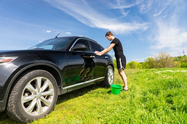 Jovem com balde verde lavando veículo preto de luxo em campo verde gramado em dia ensolarado com céu azul