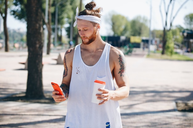 Jovem com bandana branca, vestido com uma camiseta branca, usando telefone celular e tem um copo de plástico na mão no campo de esportes do lado de fora em um dia ensolarado.