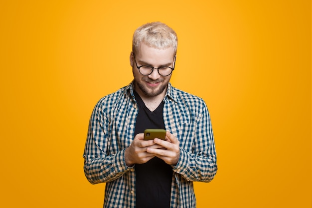 Jovem com barba e cabelo loiro usando um telefone está conversando em uma parede amarela