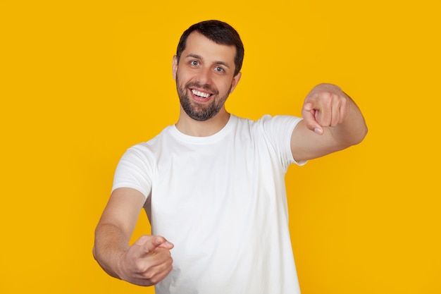 Jovem com barba em uma camiseta branca, apontando com os dedos para a câmera com uma cara feliz e engraçada.