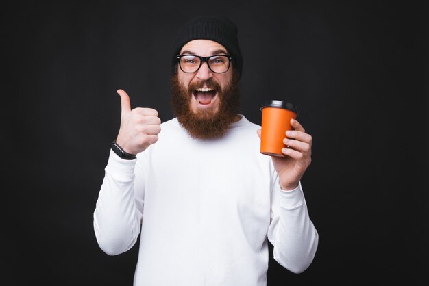 Jovem com barba está aparecendo saiu um polegar e segurando um copo com água de enxada.