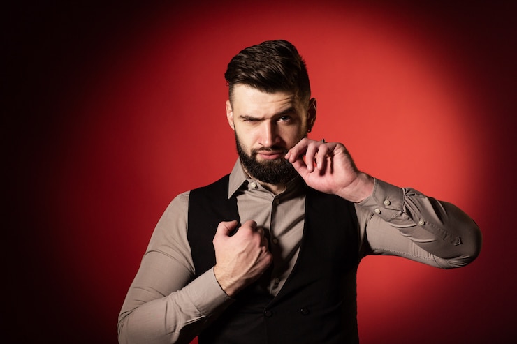 Foto jovem com barba no colete preto, posando no estúdio com fundo vermelho. um homem ajeita o bigode com a mão. fechar-se