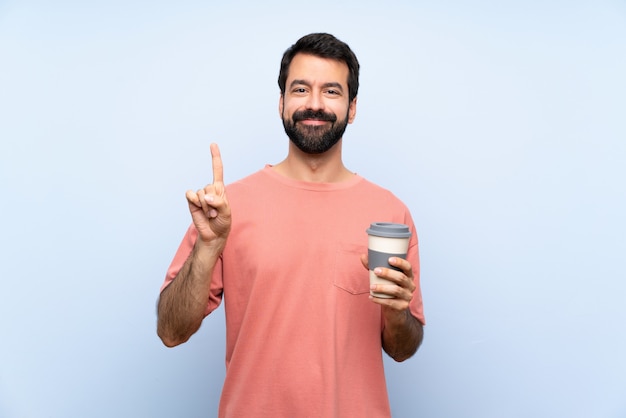 Jovem com barba segurando um café para viagem isolado parede azul mostrando e levantando um dedo em sinal dos melhores