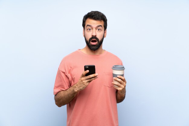 Jovem com barba segurando um café para viagem isolado parede azul surpreendeu e enviando uma mensagem