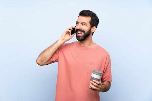 Jovem, com barba, segurando um café take away sobre parede azul isolada, mantendo uma conversa com o telefone móvel