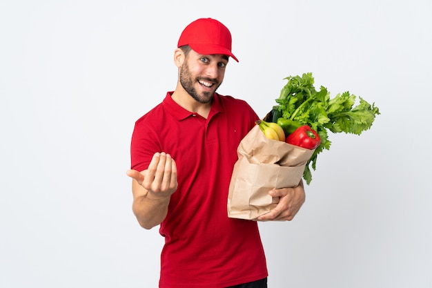 Jovem, com barba, segurando um saco cheio de legumes, isolado na parede branca, convidando para vir