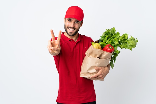 Jovem com barba segurando uma sacola cheia de vegetais isolados no fundo branco sorrindo e mostrando o sinal da vitória