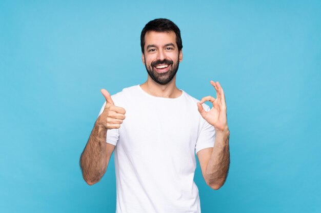 Jovem com barba sobre azul isolado mostrando sinal bem e polegar para cima gesto