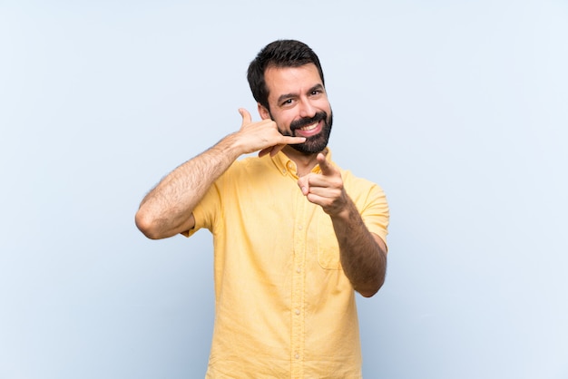 Jovem com barba sobre parede azul isolada, fazendo gesto de telefone e apontando a frente