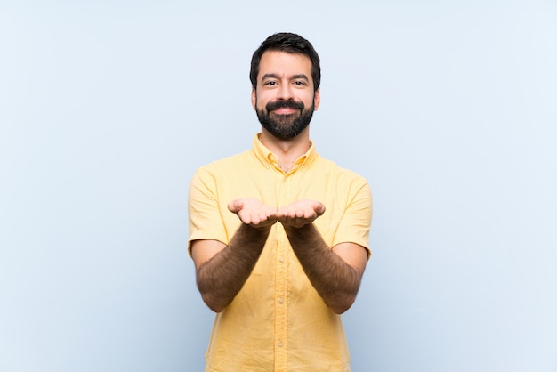 Jovem com barba sobre parede azul isolada, segurando copyspace imaginário na palma da mão para inserir um anúncio