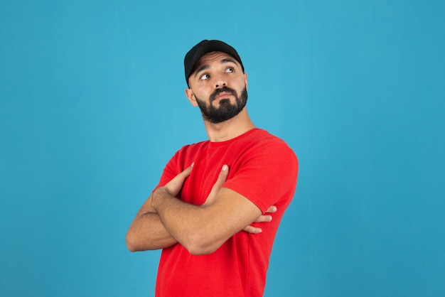 Jovem com boné vestindo camiseta vermelha em pé com os braços cruzados.