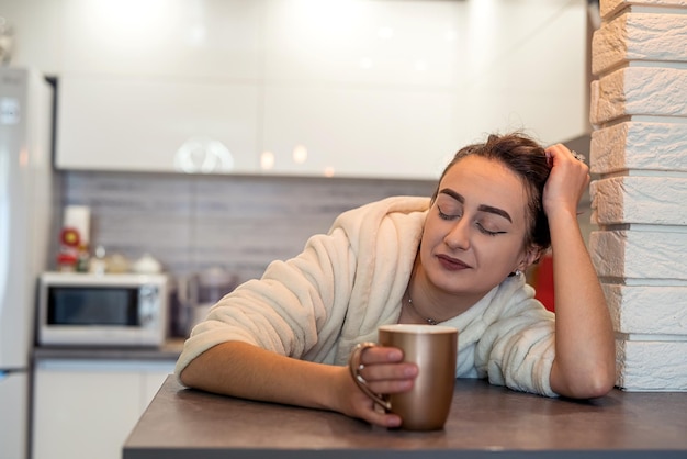 Jovem com cabelo bonito em um jaleco branco tomando café na cozinha pela manhã.