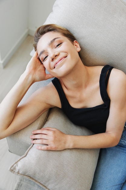 Jovem com cabelo curto se divertindo em casa no sofá sorriso e férias de felicidade em casa posando natural sem filtros espaço de cópia gratuita