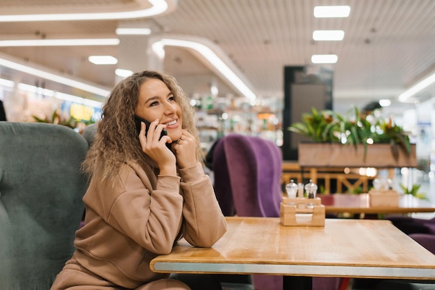 Jovem com cabelo encaracolado está sentada em uma mesa de café e conversando alegremente em um smartphone