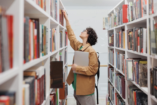 Jovem com cabelo encaracolado na biblioteca entre prateleiras com livros aluna realiza trabalho científico